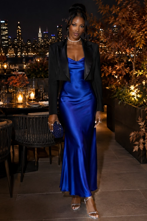 A woman wears a royal blue satin maxi dress with a cropped black blazer, silver necklace, clutch, and strappy heels at an evening rooftop setting.