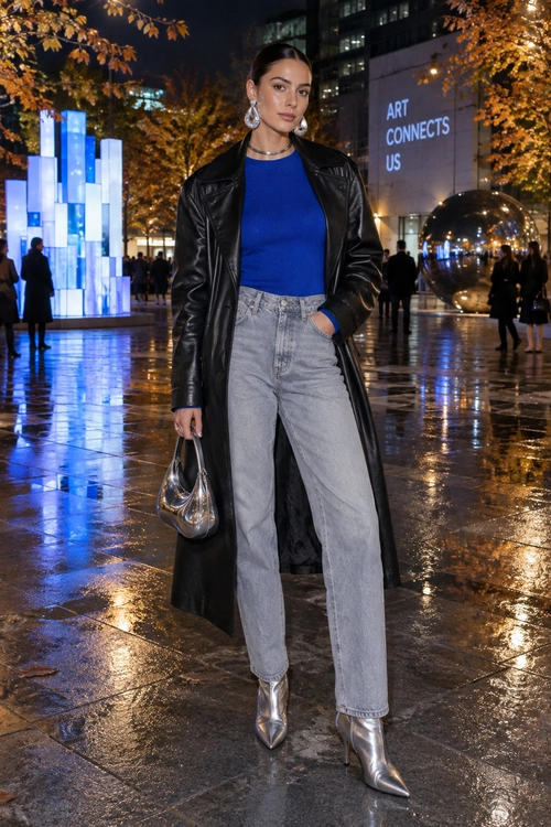 A woman wears a royal blue top, long black leather coat, gray jeans, and silver pointed ankle boots on a wet city street at night.