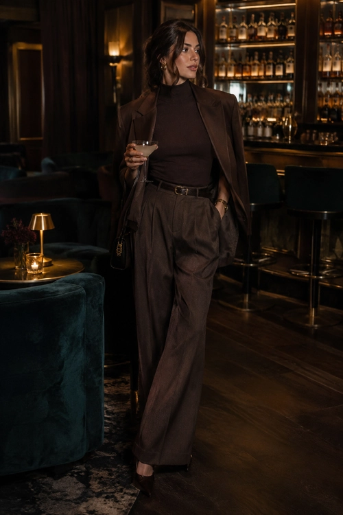 Woman wearing a dark brown blazer, high-neck top, belted wide-leg trousers, and heeled shoes in an elegant bar setting.