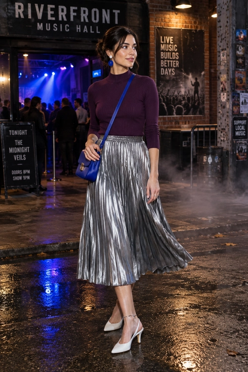 A woman wears a burgundy long-sleeve top with a metallic silver pleated midi skirt, blue crossbody bag, and pointed-toe heels outside a music hall.