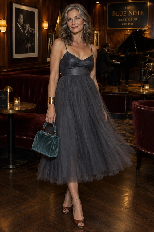 A woman wears a charcoal spaghetti-strap tulle midi dress with strappy heels, a teal handbag, and a wide cuff bracelet in a jazz club.
