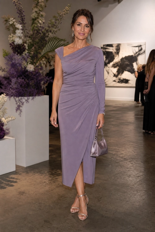 A woman wears a lavender ruched midi dress with an asymmetric neckline, metallic strappy sandals, and a matching metallic handbag.