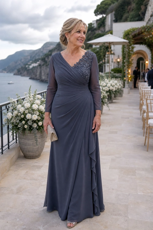 A woman wears a slate blue floor-length gown with sheer sleeves, a draped bodice, lace embellishment, and a cascading ruffle skirt.